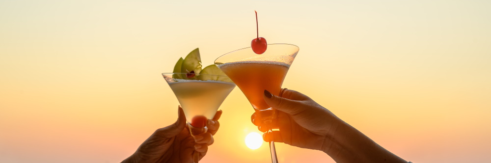 Photo of two women clinking their cocktails in front of an orange and gold sunset